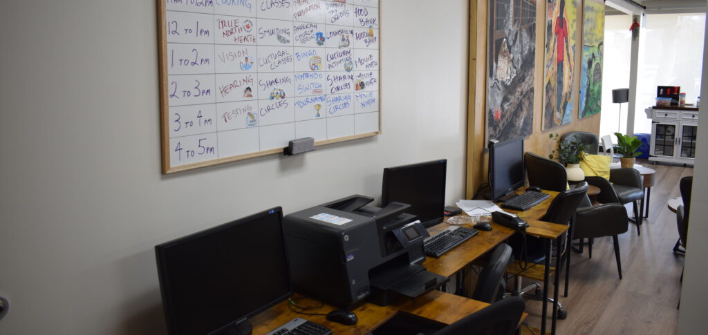 Computers and printer on table. Whiteboard on wall with schedule. Three part mural above chairs