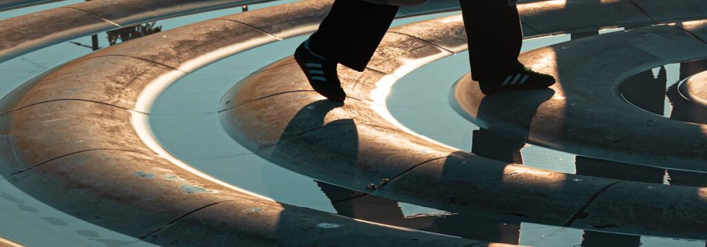 Shadowy figures cast on a spiral fountain with reflections at sunset, creating an abstract urban scene.